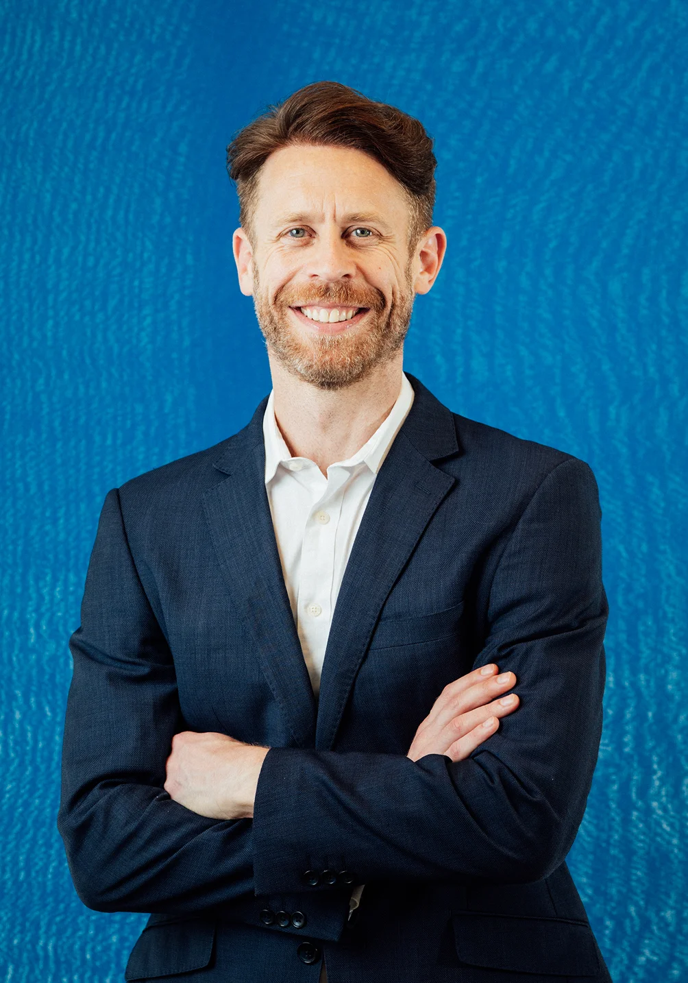 Dr Anthony Barker in navy suit smiling with arms crossed against blue background