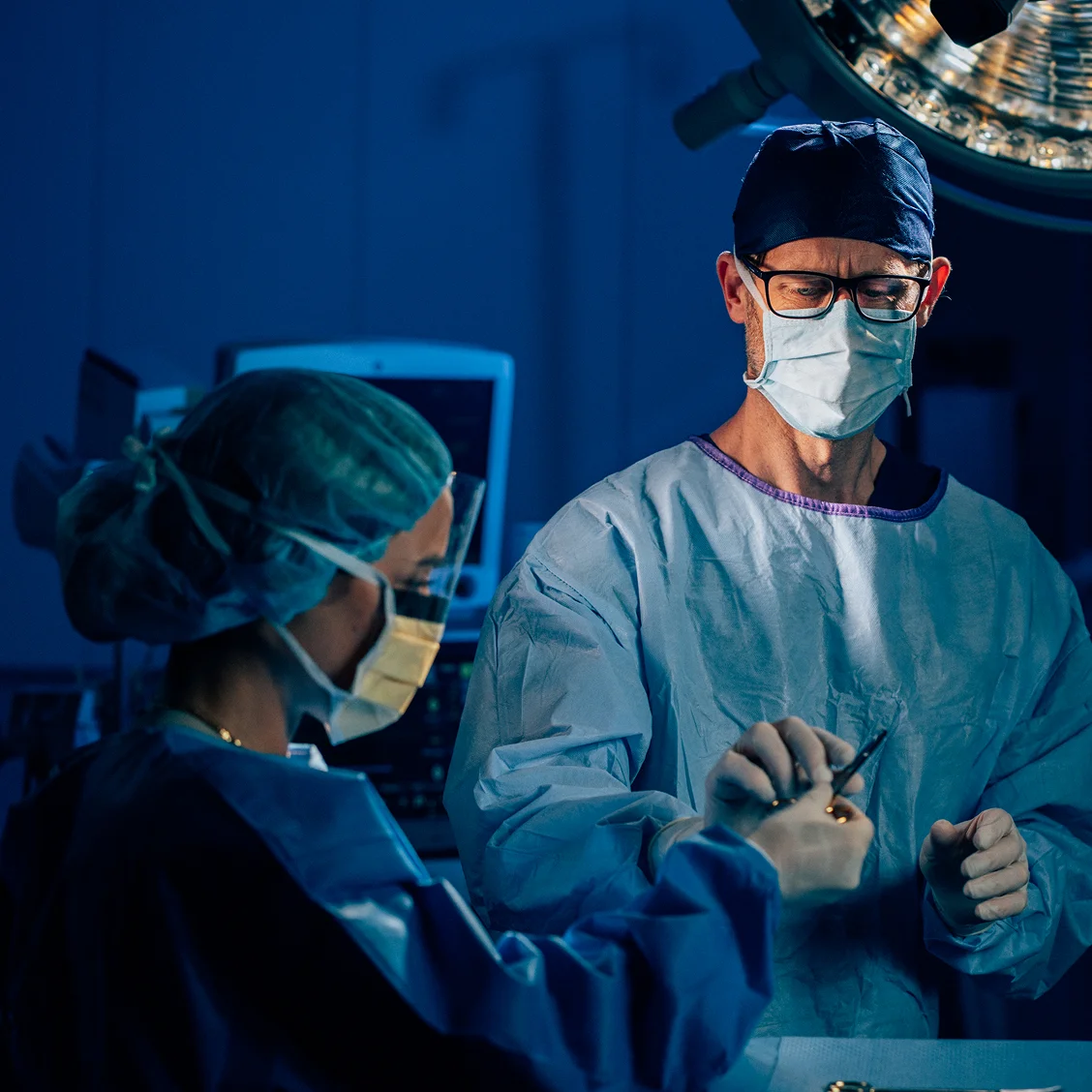 Dr Anthony Barker in masks and scrubs performing operation in hospital operating room with blue surgical lighting - Reconstuction in New South Wales, Australia