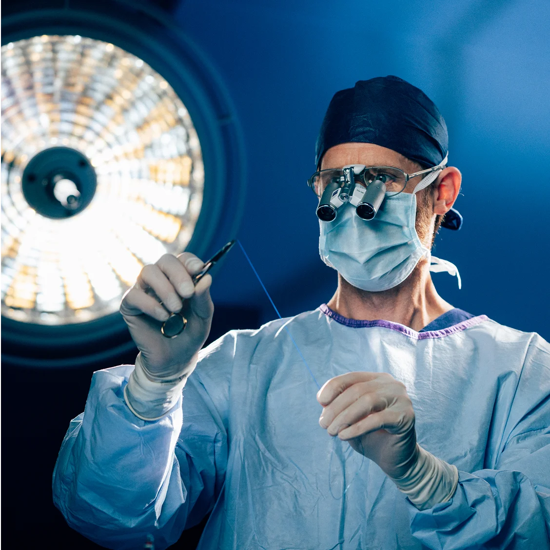 Dr Anthony Barker in blue scrubs and mask holding surgical instrument under operating room light during medical procedure - Reconstruction in New South Wales, Australia